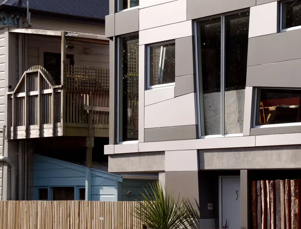 Exterior view of a contemporary multi-storey building extension or infill beside an older weatherboard dwelling. The newer facade uses large rectangular light-grey panels, dark window frames, extensive vertical glazing, and exposed concrete elements. An older timber balcony, decorative porch, weatherboard cladding, and a timber privacy fence remain visible alongside the modern addition.