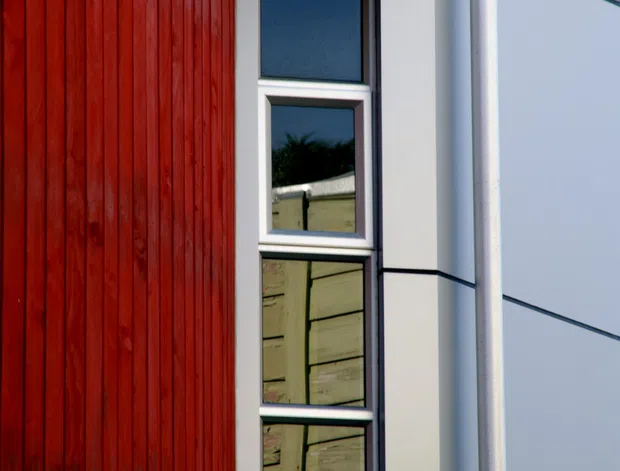 Close-up exterior view of a contemporary building facade, featuring vertical red timber cladding beside a narrow stacked window and large pale grey-blue wall panels. Dark horizontal joints, white/silver window framing, and a visible downpipe create a crisp, geometric composition.