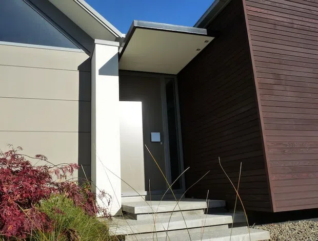 Contemporary residential entry viewed from outdoors, featuring a recessed dark front door beneath a broad flat canopy, pale horizontal wall cladding, a projecting white structural volume, and a tall dark timber-clad facade. Concrete steps lead through landscaped planting and gravel toward the entrance under a clear blue sky.