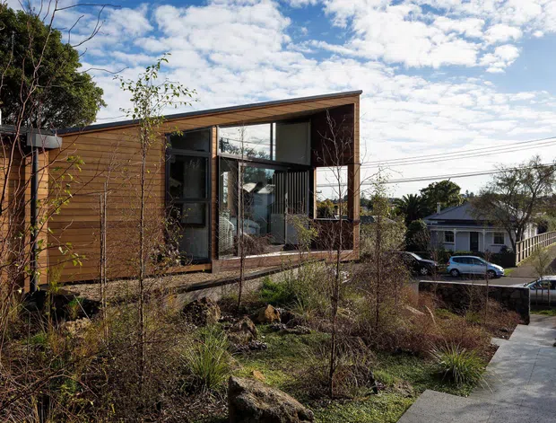 A compact contemporary single-storey house on a sloping suburban site, viewed from the landscaped front path. The building has warm horizontal timber cladding, a low-pitched roof, dark-framed floor-to-ceiling glazing, and a projecting glazed living area. Native-style planting and rockwork surround the entry, with neighbouring suburban homes and overhead utility lines visible in the background.