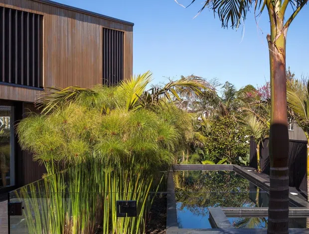 Contemporary two-storey home exterior with dark vertical timber-like cladding, tall screened windows, and extensive tropical landscaping surrounding a reflective pool or water feature. A glazed balustrade and outdoor walkway frame the foreground, with palms and ornamental grasses creating a lush, private setting under clear skies.