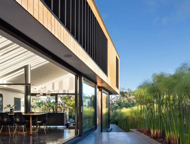 Exterior view of a contemporary two-storey residence with a projecting upper volume clad in warm vertical timber and dark vertical screening. Full-height glazing opens onto a paved terrace, revealing a bright open-plan dining and living interior with black dining chairs, pendant lighting, and a kitchen island. Lush planted landscaping borders the terrace under a clear blue sky.