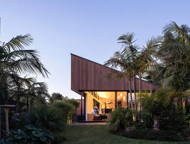 Exterior dusk view of a contemporary elevated house surrounded by dense tropical landscaping. The home features a dramatic asymmetrical pitched roof clad in dark vertical timber, extensive warm-lit glazing, slender supports, and a landscaped grass approach framed by palms and lush planting.
