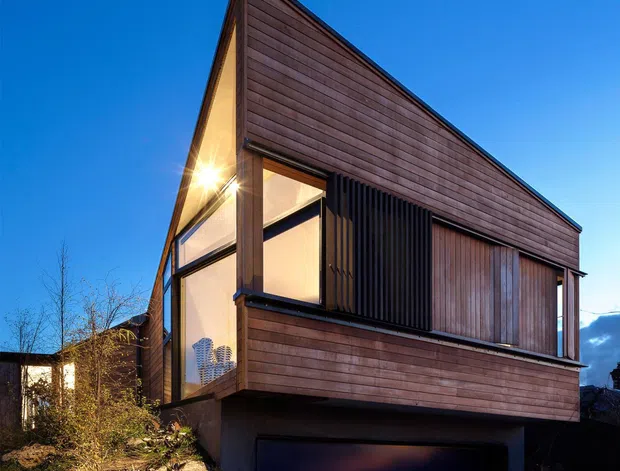 Exterior dusk photograph of a contemporary, angular timber-clad house on a rocky sloping site. The compact two-storey dwelling has a steep asymmetrical gable-like roof form, extensive dark-framed glazing, vertical timber screening, projecting upper-level volume, and an integrated dark garage beneath. Native-style landscaped planting and exposed rocks surround the building.
