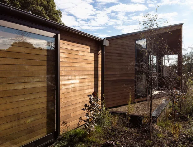 Exterior view of a low-rise contemporary building set within a planted garden. The facade is clad in horizontal warm timber boards, with dark-framed glazing and an angular mono-pitched roofline. An irregular natural-stone path leads through the landscaped foreground, and a covered or glazed connection appears between the timber-clad volumes. Bright daylight and a partly cloudy sky emphasize the warm, natural character of the architecture.