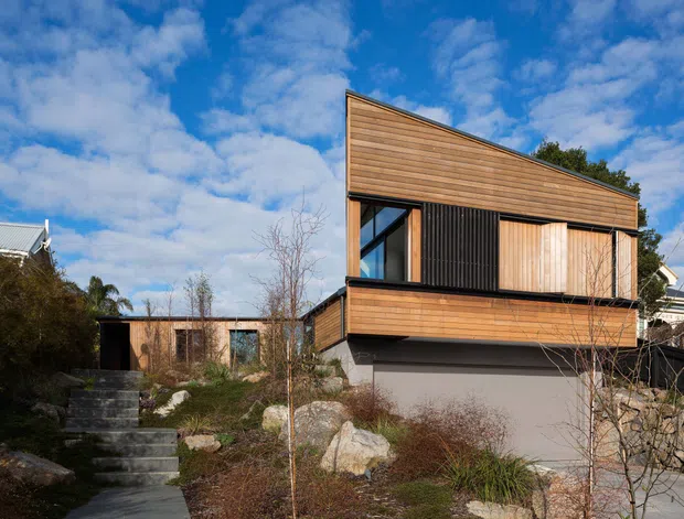 Exterior view of a contemporary two-storey home on a landscaped, rocky sloped site. The building has warm horizontal timber cladding, a sharply angled single-pitched roof, dark vertical screening, large windows, and a raised upper level above a grey lower storey. Stone retaining walls, concrete steps, and native-style planting frame the approach.