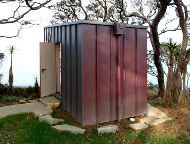 A compact standalone rectangular structure set among coastal vegetation, clad in dark ribbed metal panels with a rust-red finish. A simple hinged door opens onto a rough stone-paved path, with exposed stone edging and a small flat roof. The surrounding trees and grasses give the scene a natural, secluded setting.