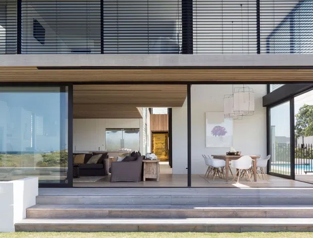 Contemporary multi-level residential home viewed from the garden, with broad floor-to-ceiling glazing opening onto a stepped terrace. The visible interior combines a living room and dining area, while the upper level features horizontal louvres and a projecting timber-lined soffit. A swimming pool is visible to the right, and the facade uses dark-framed glazing, pale rendered walls and warm timber detailing.