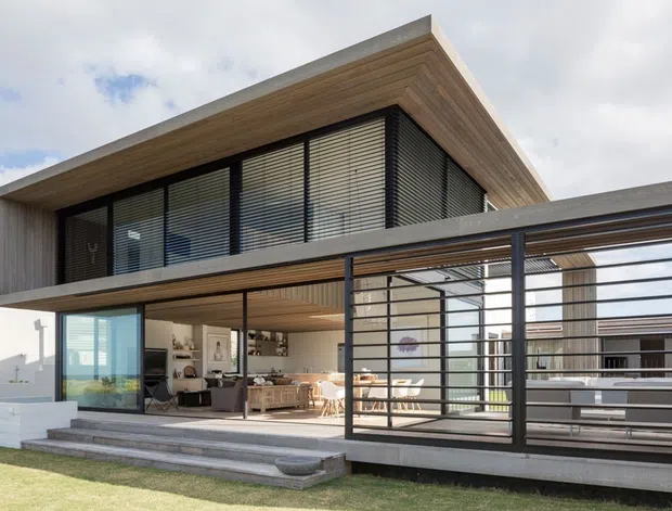 Exterior view of a contemporary two-storey residence with a projecting flat roof, warm timber soffits and cladding, extensive black-framed glazing, and horizontal louvres. The open-plan living area is visible through the ground-floor glass, with a covered outdoor terrace enclosed by a black metal screen structure and steps leading to a lawn.
