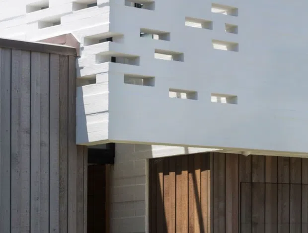 Close-up exterior view of a minimalist modern building featuring a white rendered or painted masonry facade with a regular pattern of narrow recessed openings. A projecting upper volume sits above a darker timber-clad entrance or garage-like opening, creating strong geometric contrast between white masonry and vertical brown timber boards.