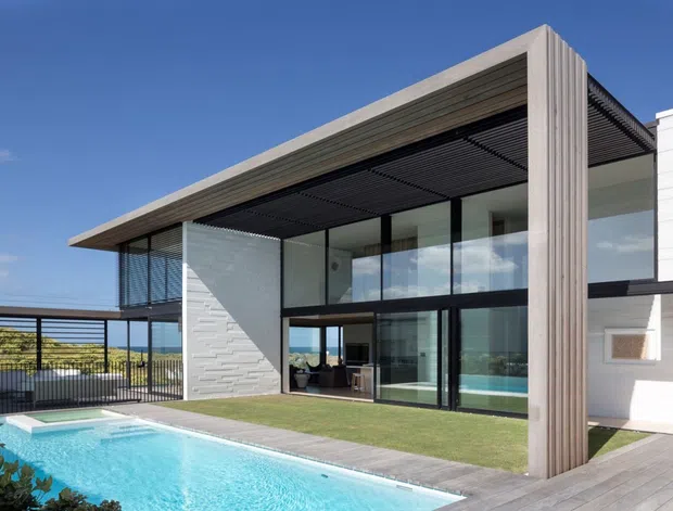 Modern two-storey residential exterior with a strong rectangular portal frame, extensive floor-to-ceiling glazing, dark horizontal sun-shading screens, pale textured masonry walls, and a timber-toned vertical feature frame. A swimming pool, lawn, and paved terrace occupy the foreground, with an outdoor covered entertaining area visible to the left.