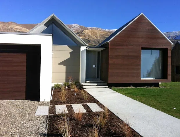 Exterior view of a contemporary single-storey residential building composed of connected gabled volumes. The facade combines dark horizontal timber cladding with pale horizontal weatherboard-style cladding, large dark-framed glazing, and a prominent timber garage door. A simple concrete path crosses a landscaped front garden with gravel and ornamental grasses, with alpine mountains visible in the background.