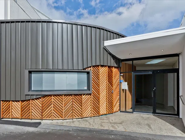 Exterior view of a contemporary building entrance with a curved dark metal-clad facade, projecting white canopy, extensive glazing, and warm timber chevron screening at the lower facade. The composition emphasizes the main entry and contrasting facade materials beneath a partly cloudy sky.