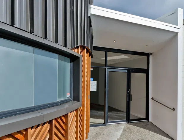 Exterior view of a contemporary building entrance, featuring a projecting white canopy, black-framed glazed double doors, a large horizontal window, vertically ribbed dark metal cladding, and decorative timber lattice screening. A concrete access path leads to the entry, with a handrail along the white wall.