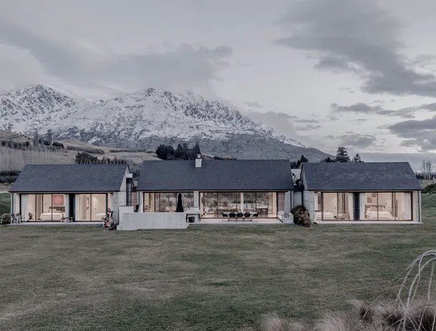 A low, single-storey contemporary residence arranged as three connected pavilion-like volumes across a broad grassed site. The home has dark pitched roofs, extensive floor-to-ceiling glazing, pale exterior walls, and a central glazed living area. Snow-covered alpine mountains form a prominent backdrop, with landscaped planting and open rural grounds surrounding the house. The scene is photographed in subdued, overcast light.