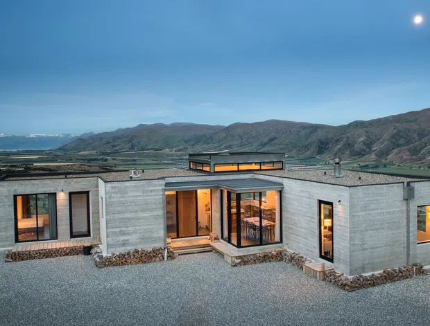 A low-profile modern concrete house set in a rugged mountainous landscape, photographed at dusk. The single-storey dwelling has a rectilinear, courtyard-like plan with exposed grey concrete walls, extensive dark-framed glazing, warm interior lighting, a raised central roof element or clerestory, and gravel landscaping with stone-edged planting beds. Mountains and a broad rural valley form the background, with the moon visible in the blue evening sky.