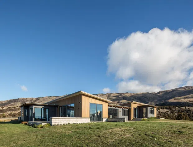 A low-profile contemporary single-storey home set in an expansive rural landscape, with warm vertical timber cladding, dark window and door frames, broad glazing, and a shallow pitched roof. The house sits on a flat grassy site backed by dry, rugged hills beneath a clear blue sky.