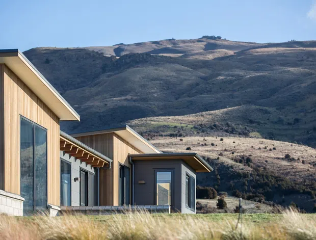 Exterior view of a contemporary single-storey residence composed of several connected timber-clad volumes, with large glazed openings and low-pitched projecting roofs. The home sits in an open, rugged mountain landscape with dry grasses in the foreground and broad hills behind it.