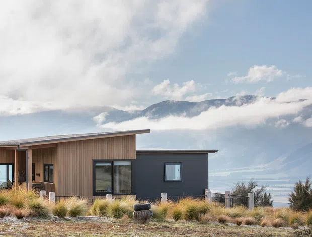 Single-storey contemporary house on a rugged elevated site, featuring dark vertical cladding, warm timber-clad sections, broad roof overhangs, and large glazed openings. Ornamental grasses and low planting surround the building, with dramatic cloud-covered mountains and a distant body of water visible behind it.
