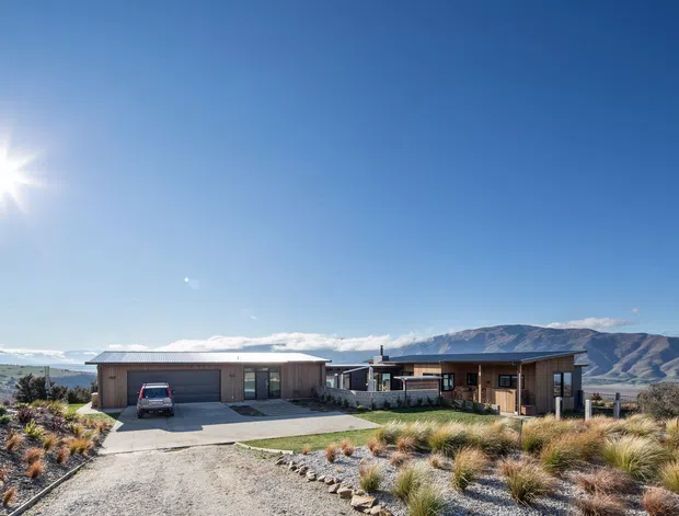 A wide exterior photograph of a low-profile contemporary house set in an open, dry landscaped site. The home has a long horizontal form, muted dark cladding, a prominent garage, large glazed openings, and a partially enclosed courtyard or terrace. Ornamental grasses and gravel surround the driveway, with expansive mountains visible in the background under a clear blue sky.