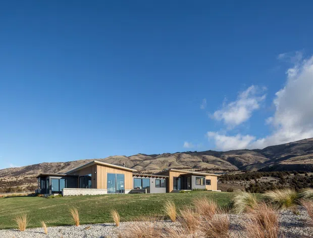 A low-profile contemporary single-storey house set in a broad, landscaped rural or alpine-like setting. The building has a long horizontal composition with timber-clad sections, dark-framed glazing, flat or very low-pitched roof forms, and a central recessed entry or courtyard-like break. A manicured lawn transitions to gravel and ornamental grasses in the foreground, with dry rolling hills and a clear blue sky beyond.
