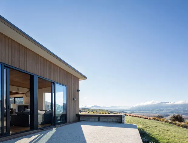 Exterior view of a contemporary single-storey home set on an elevated, open landscape. The building has vertical timber cladding, dark-framed floor-to-ceiling glazing, and a low-pitched roof with broad eaves. A paved terrace and low retaining wall overlook a broad valley and distant mountains. Interior living furnishings are visible through the glazing.