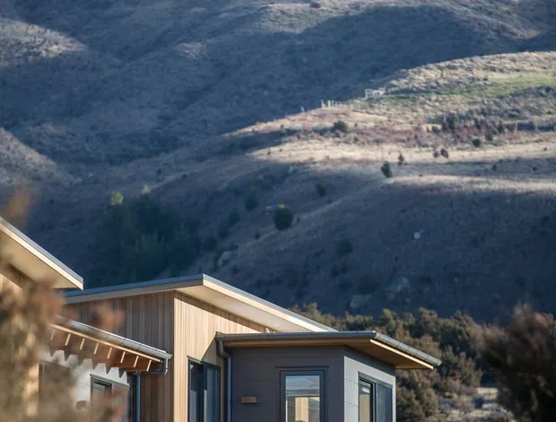 Exterior view of a compact contemporary house set in a rugged mountainous landscape. The building features low-pitched intersecting roof planes, vertical timber-clad walls, dark horizontal cladding, large windows, and a sheltered entry or outdoor terrace. Native grasses and scrub surround the house, with steep dry hills forming the backdrop.