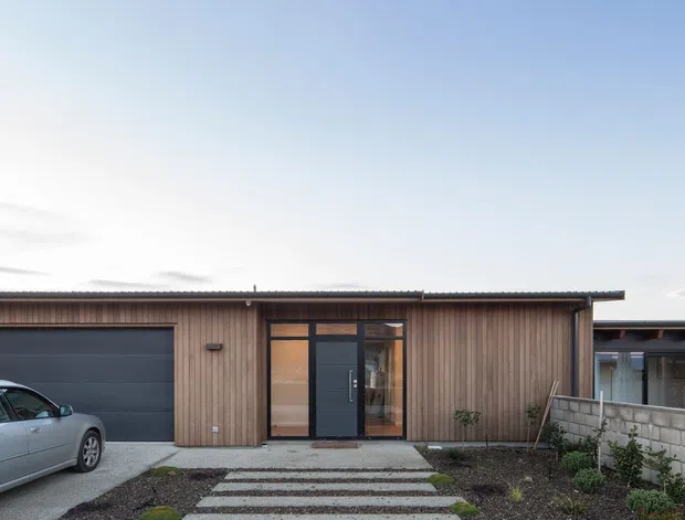 Front exterior view of a low, single-storey residence with a simple flat or very low-pitched roof, vertical timber cladding, dark grey garage door and entrance door, and glazed entry surround. A gravelled front garden with rectangular concrete stepping slabs forms the pathway to the entrance, with a concrete driveway and partial carport or adjoining structure visible at the side.