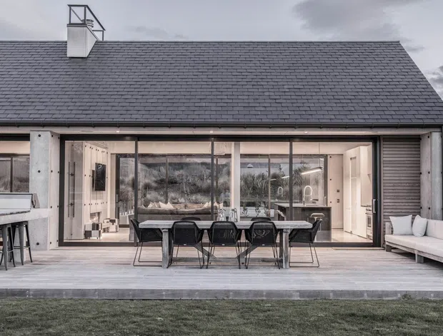 Rear exterior view of a contemporary single-storey home with a broad dark pitched roof, pale rendered walls, extensive floor-to-ceiling glazing, and large sliding doors opening onto a timber deck. The deck includes an outdoor dining setting, built-in outdoor kitchen or barbecue area, and a pale outdoor lounge. A bright open-plan interior with kitchen cabinetry is visible through the glazing.