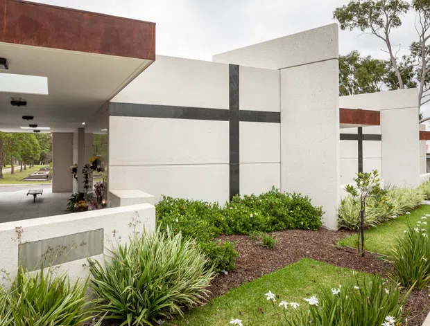 Exterior view of a contemporary, low-slung building with crisp white rendered walls, dark vertical and horizontal feature bands, and rust-toned metal roof or fascia elements. A covered entry or driveway passage is visible on the left, opening toward a landscaped garden with ornamental grasses, shrubs, and small trees. The composition emphasizes strong geometric planes, deep roof overhangs, and a restrained modern material palette.
