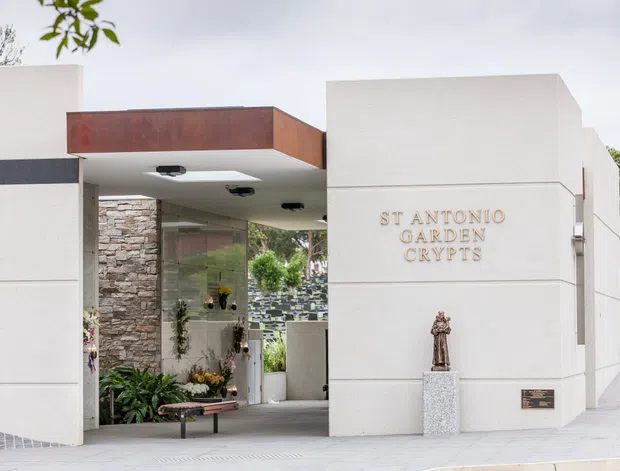 Exterior view of St Antonio Garden Crypts, featuring a minimalist white rendered entrance structure with clean geometric forms, a rust-toned metal canopy, stone-clad wall, glazed entry area, landscaped planting, and a paved forecourt. The building signage and religious statue indicate a memorial or cemetery facility.