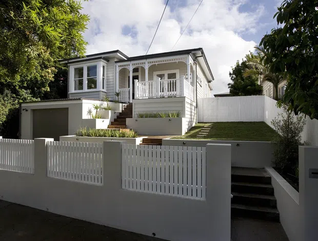 A street-facing exterior view of a raised, light-painted timber house with a front verandah, decorative fretwork, bay window, garage, stepped entry, manicured lawn, and white picket-style boundary fencing. The home has a traditional bungalow character with a simple gabled roof and strong white-and-grey detailing.