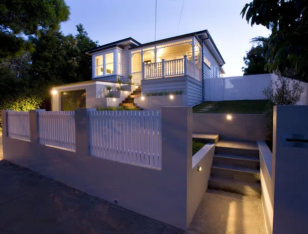Twilight exterior view of a raised, multi-level suburban house behind a rendered boundary wall and vertical slatted gates. The home features light weatherboard cladding, a pitched gabled roof, illuminated windows, a covered balcony with timber-style balustrade, exterior stair access, landscaped planting, and integrated wall lighting.
