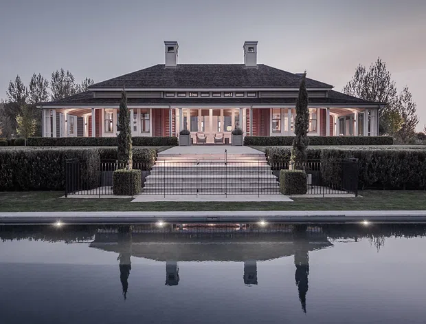 Symmetrical exterior view of a formal, single-storey pavilion-style residence with a broad low-pitched hipped roof, twin chimneys, classical columns and detailing, and a centered staircase leading through manicured hedges to a long reflecting pool. The subdued dusk lighting and still water create a refined, dramatic atmosphere.