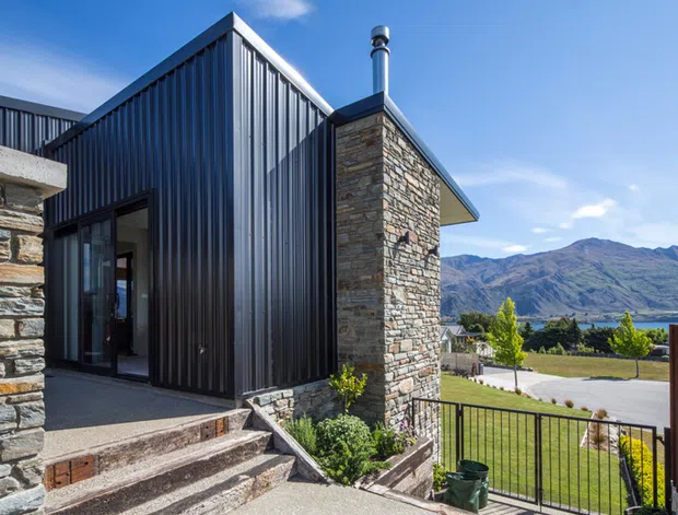 Exterior view of a contemporary single-storey home featuring dark vertical metal cladding, natural stacked-stone walls, a pitched roof form, and a glazed entry. The property sits on a landscaped, elevated site with views toward a mountain range and lake or valley beyond.