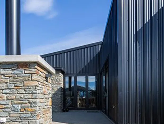 Exterior view of a contemporary building entrance reached by concrete steps. The facade combines dark vertically ribbed metal cladding with a low wall of layered natural stone, dark-framed glazing, and a prominent cylindrical metal chimney or flue. Bright blue sky and strong sunlight emphasize the crisp geometric forms and contrasting materials.