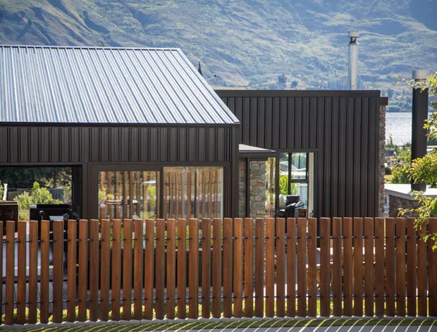 Exterior view of a contemporary dark-clad house with a steep corrugated metal gable roof, large glazed openings, timber fencing, and a chimney. The home sits in a mountainous setting with a glimpse of water beyond.