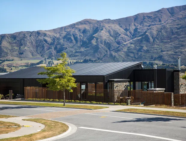 Contemporary low-rise building with dark corrugated metal roofing and cladding, stone masonry walls, timber fencing, and glazed openings, viewed from a suburban street. Rugged mountain ranges and a rural valley form the background.