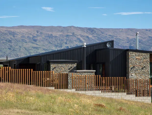 Contemporary single-storey residence on a sloping site, featuring dark vertical metal cladding, extensive natural stone veneer, timber-framed glazing, and a low angular pitched roof. A timber slat fence and stone retaining wall define the foreground, with rugged mountains forming the distant backdrop.