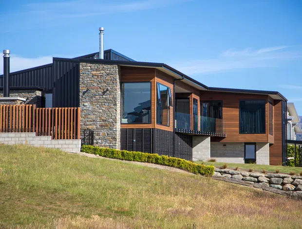 Exterior view of a contemporary multi-level hillside home with dark metal cladding, warm timber wall sections, extensive natural stonework, large glazed openings, and a low-pitched asymmetrical roof. The house is set within a landscaped sloping site with a timber privacy screen, retaining wall, and mountain scenery in the distance.