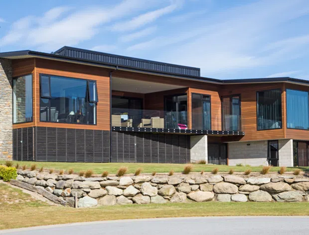 Contemporary single-storey residential home with a strongly horizontal composition, flat rooflines, warm timber cladding, dark metal screening and extensive blue-tinted glazing. A prominent stacked-stone wall forms the left end and the raised front facade is supported above a landscaped rock retaining wall. The central recessed balcony or outdoor sitting area is visible behind glazed doors, with a suburban streetscape beyond.