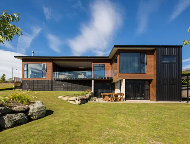 Contemporary two-storey residence with a strong horizontal composition, dark vertical cladding, warm timber wall sections, expansive glazing, and a covered upper-level terrace. The house sits within a landscaped lawn with natural stone edging and a partially visible carport to the right.