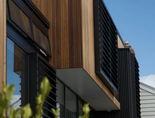 Exterior architectural photograph of a contemporary multi-level building featuring warm vertical timber cladding, dark charcoal metal framing and vertical screening, projecting balcony or canopy elements, and extensive glazing. A neighbouring light grey weatherboard structure is partially visible to the right, with landscaped planting in the foreground and blue sky behind.