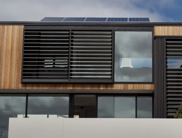 Contemporary multi-level residential facade with warm vertical timber cladding, dark metal louvred screens, expansive glazing, a flat roof, and rooftop solar panels. A white rendered boundary wall and neighbouring weatherboard structure are visible at the lower and right edges.
