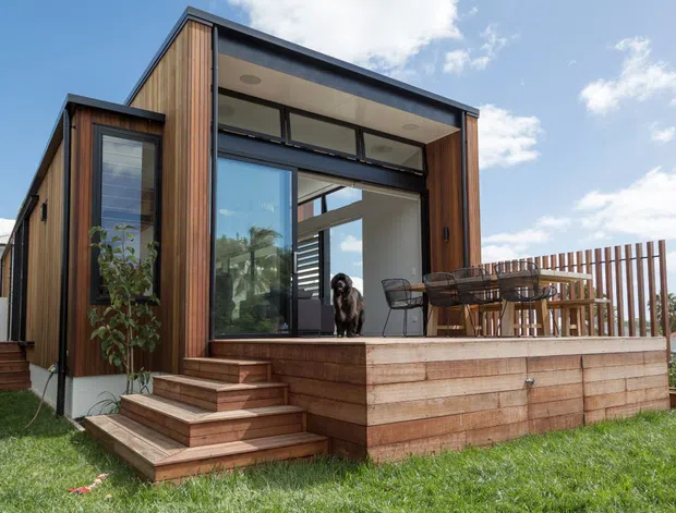 Exterior view of a compact modern timber-clad home or extension with a raised timber deck, outdoor dining area, large glazed opening, and broad roof overhang. Vertical timber battens form a privacy screen along the deck, with steps leading down to a lawn.