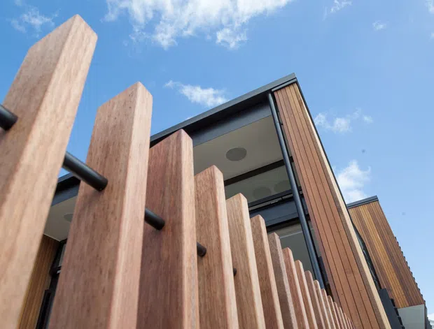 Low-angle exterior view of a contemporary multi-level building with prominent vertical timber screening, dark horizontal rails, timber-clad structural elements, large glazed openings, and projecting balconies beneath a dark roof edge. The image is set against a bright blue sky.