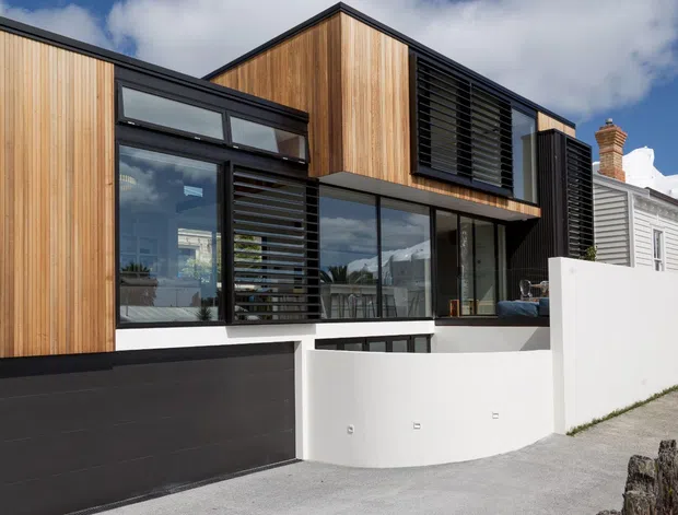 Street-facing exterior of a contemporary multi-level house, combining warm vertical timber cladding with dark metal-framed glazing, black louvred screens, and a white rendered boundary wall. The design sits alongside a traditional weatherboard house, with a pitched roof form, brick chimney, and urban residential streetscape visible.