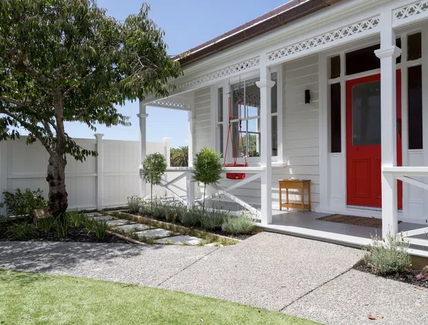 Bright exterior view of a white weatherboard home with a covered front veranda, ornate decorative fretwork, white posts and railings, and a striking red front door. A textured concrete path leads through a landscaped front garden with lawn, shrubs, and a mature tree. The facade has a traditional bungalow character with neatly detailed trim and a low-pitched roof.