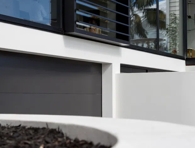 Close-up exterior architectural photograph of a contemporary multi-level residence. The facade features white rendered walls, dark charcoal garage and window frames, large glazed openings, and prominent horizontal black louvres for solar shading. A curved white rendered planter wall occupies the foreground, with palm vegetation and a neighbouring light-clad building reflected in the glazing.