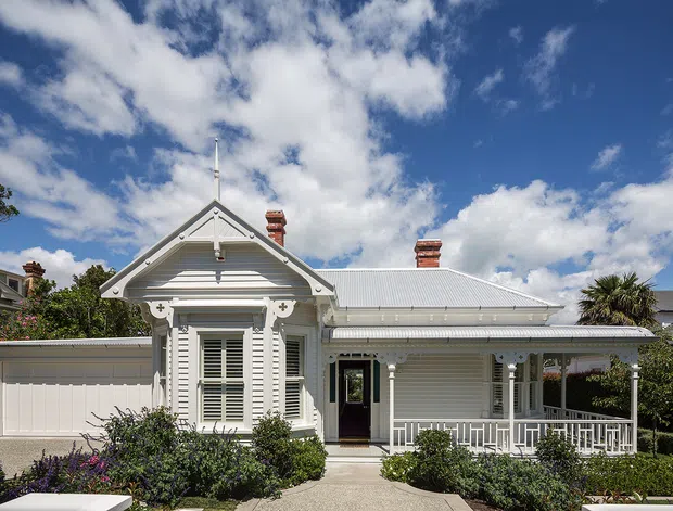Front exterior view of a renovated single-storey white weatherboard house with a prominent gabled bay, decorative Victorian-style trim, corrugated metal roof, front veranda, enclosed garage, chimney stacks, and landscaped garden under a bright blue sky.